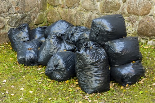 A collection of several large black plastic rubbish bags, tightly sealed and piled together on a grassy area near a stone wall. The bags appear to contain waste materials, and they are slightly reflective due to the plastic finish. The green grass beneath the bags has scattered leaves and small stones, indicating an outdoor setting, possibly a driveway or garden area in Hendon. The stone wall in the background is composed of irregularly shaped rocks with moss or dirt accumulation, providing a natural and textured backdrop. The scene is well-lit with natural daylight, emphasizing the contrast between the dark bags and the lighter environment, illustrating typical rubbish removal and clearance activities that [COMPANY_NAME], such as Office Clearance Hendon, handle in the local area as part of their rubbish collection services.