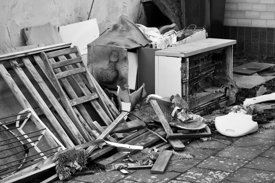 The image depicts an outdoor scene showing various discarded items piled against a wall, indicative of a rubbish clearance situation. In the foreground, there are two wooden garden chairs with slatted