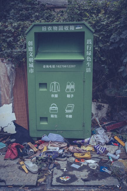 A green, outdoor clothing collection bin positioned against a backdrop of dense foliage and trees; the bin has Chinese characters on the top and sides, indicating it is used for old clothing recycling