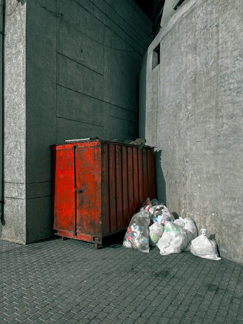 A green, outdoor clothing collection bin positioned against a backdrop of dense foliage and trees; the bin has Chinese characters on the top and sides, indicating it is used for old clothing recycling