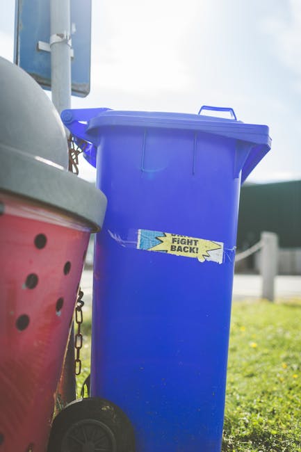 A row of black wheelie bins with closed lids line the edge of a pavement along a residential street, positioned in front of a brick building with multiple white-framed windows. A woman dressed in a da