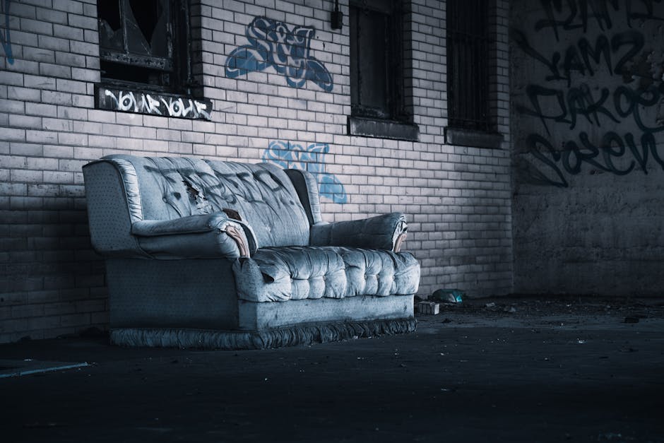 A vintage brown leather sofa with a slightly worn texture positioned against a white-painted wall inside a room. Behind the sofa, a large window divided into six panes reveals an exterior view of gree