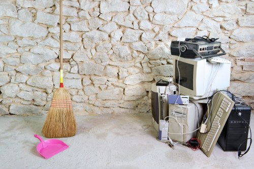 A collection of discarded household items including an old CRT computer monitor with a beige casing and a black top, a tangled bundle of cables and wires resting on and around the monitor, a small portable desktop speaker, a grey plastic-framed keyboard leaning against other objects, and a black computer tower situated on the floor near a beige mini tower case, all placed against a textured stone wall with a light-colored concrete or cement floor. Nearby, a pink plastic dustpan with a short handle and a small dust broom with natural straw bristles and a red and yellow handle are positioned on the ground, indicating a cleaning or clearing process in progress. The setting appears to be a garage, basement, or storage area, and the scene is connected to rubbish removal services in Hendon, as offered by Office Clearance Hendon, focusing on waste disposal and clearance of electronic and household clutter.
