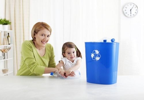 A woman with short blonde hair and a light green top is smiling and sitting at a white dining or kitchen table, accompanied by a young girl with brown hair in pigtails wearing a white shirt, both appearing pleased and engaged. In front of them on the table, there is a transparent plastic bottle. To the right, a large blue recycling bin with a white recycling symbol on its front is visible, placed on a light-colored surface. The background features a room with white curtains, a white shelving unit with decorative items, and a wall-mounted clock showing approximately 12:05. The environment suggests an indoor household setting, emphasizing waste separation and recycling, relevant to rubbish removal services offered by companies like Office Clearance Hendon, serving areas such as Hendon and surrounding London districts.