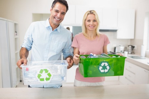 A middle-aged man and woman stand side by side in a bright, modern kitchen, each holding a clear plastic recycling bin with a green recycling symbol on the front. The man, on the left, is wearing a light blue button-up shirt with rolled-up sleeves, and he holds the bin with both hands, which appears to be filled with plastic bottles and packaging. The woman, on the right, is dressed in a soft pink T-shirt and holds a slightly larger, solid green bin containing various recyclable materials. Behind them, the kitchen features white cabinets, a stainless steel sink, and countertops, with some kitchen utensils and appliances gently visible in the background. The scene is well-lit with natural or indoor lighting, creating a warm and clean atmosphere that subtly emphasizes the importance of waste management and recycling practices, aligning with professional rubbish removal themes for a local service such as Office Clearance Hendon. The setting suggests a focus on household or office waste sorting and recycling efforts, subtly supporting local SEO signals for Hendon and surrounding areas.
